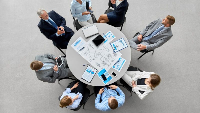 Vogelperspektive auf eine Gruppe von Menschen in formalen Anzügen an einem runden Tisch sitzend. Darauf sind Planungsmaterialien erkennbar. Der Hintergrund ist ein grauer Boden.