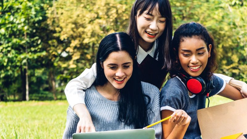 Drei junge Frauen sitzen in einem Park und arbeiten an einem Laptop.