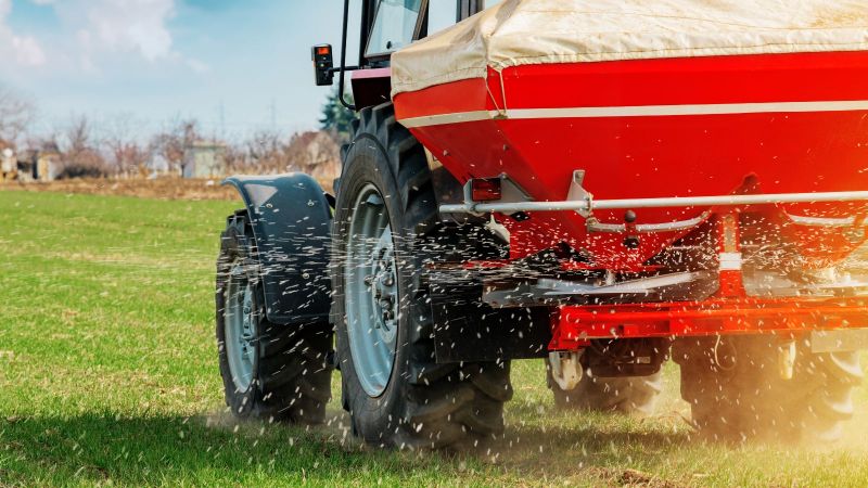 Ein Tracktor verteilt Düngemittel mithilfe seines angekoppelten Schleuderstreuers auf einer stoppeligen Rasenfläche.